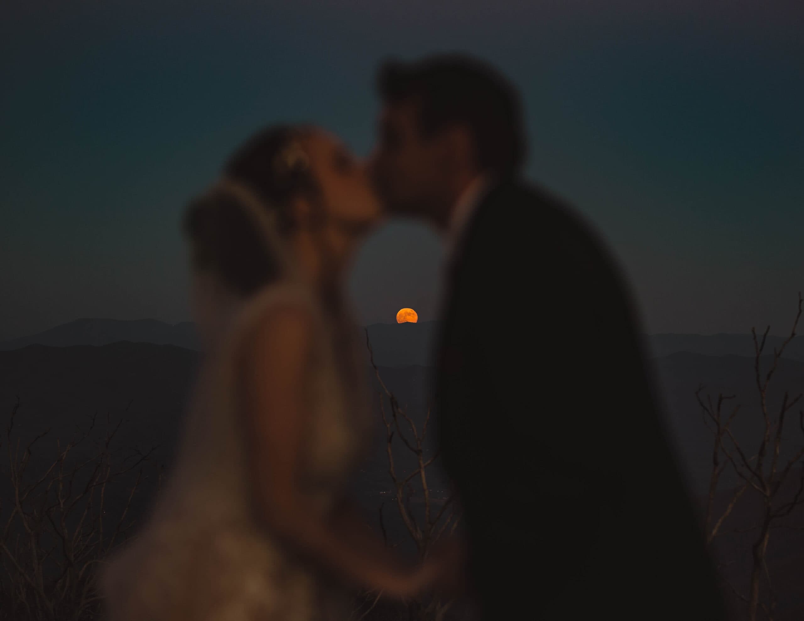 A bride and groom kissing with the moon and mountains in the background at dusk for their North Georgia elopement.