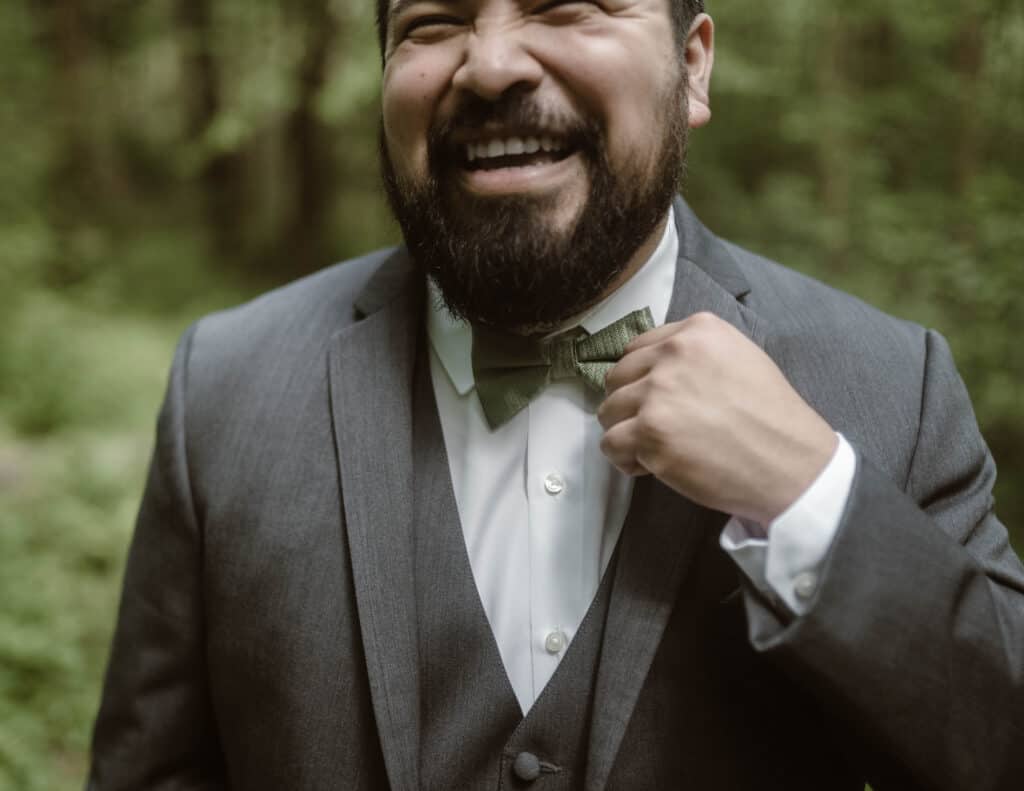 A groom smiling and laughing while holding his bow tie. 