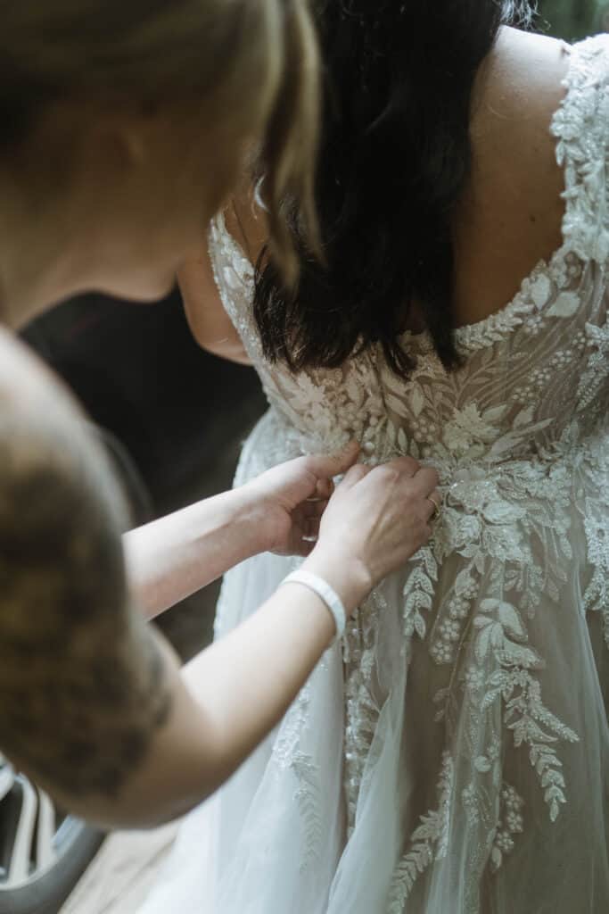 A mother-of-the-bride helping the bride button her dress before their waterfall ceremony. 