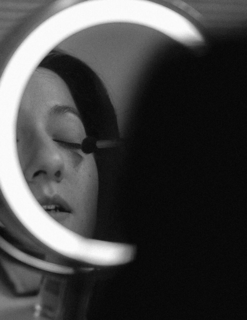 A black and white image depicting a girl applying eye makeup in a mirror. 