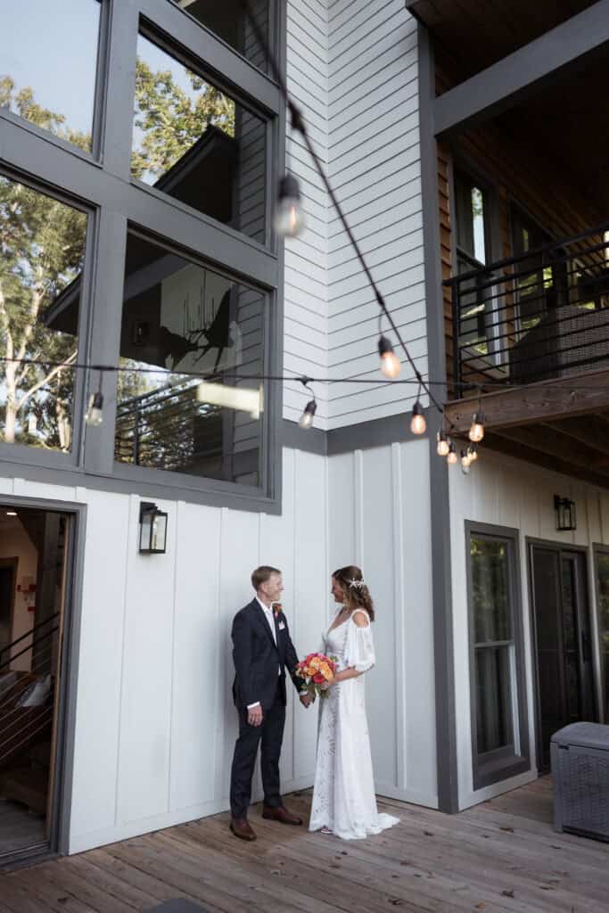 A newlywed couple look lovingly at teach other while holding hands on a large porch with hanging lights. 