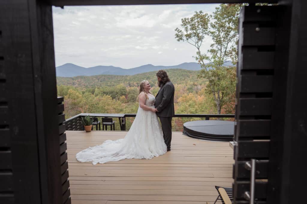 A newlywed couple holding hands facing each other while standing on the porch of a cabin overlooking the mountains. 