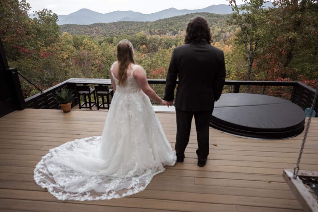 A newlywed couple standing hand in hand looking at the mountain view from the porch of their cabin rental. 