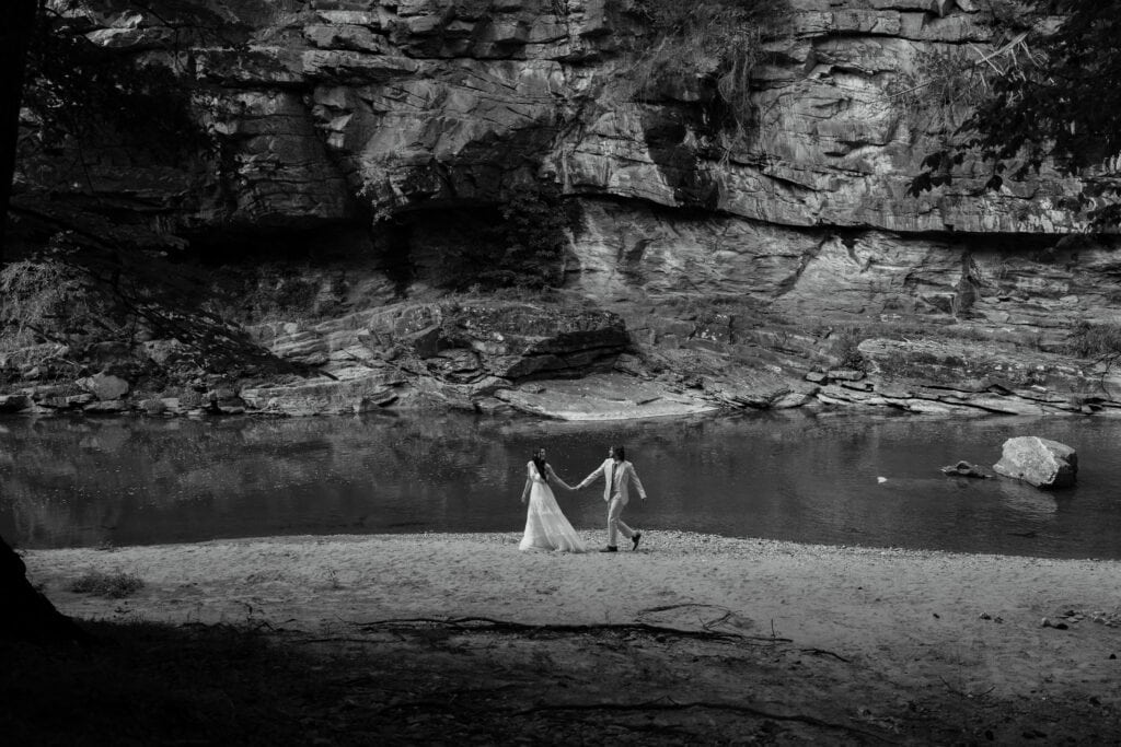 A couple walking hand-in-hand on the sand with a river and rock face behind them. 