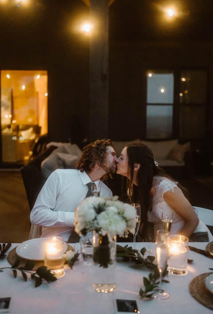 A newlywed couple kiss with hanging lights while waiting for their first meal as a married couple. 