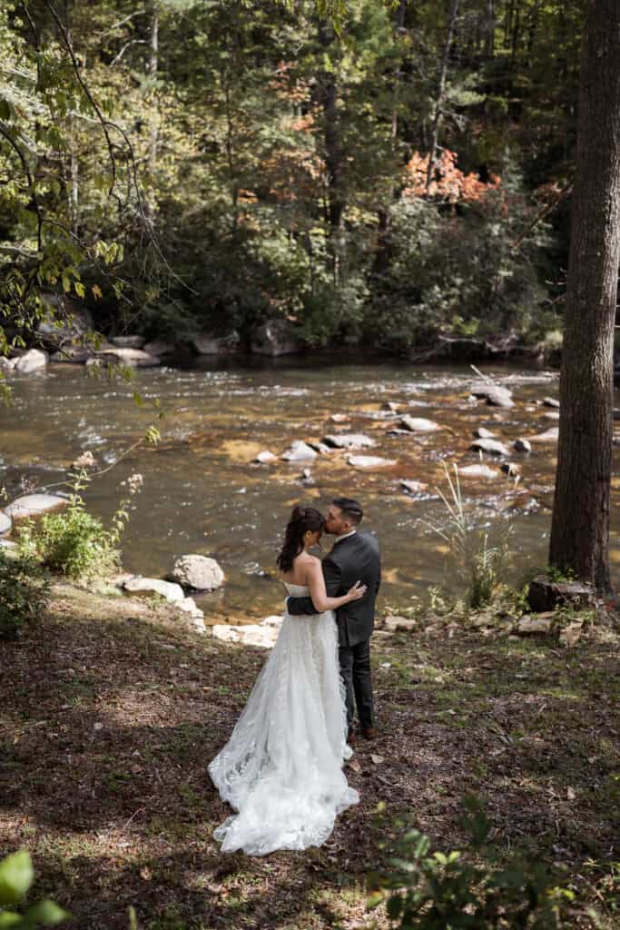 A newlywed couple standing by a river kissing each other's foreheads. 