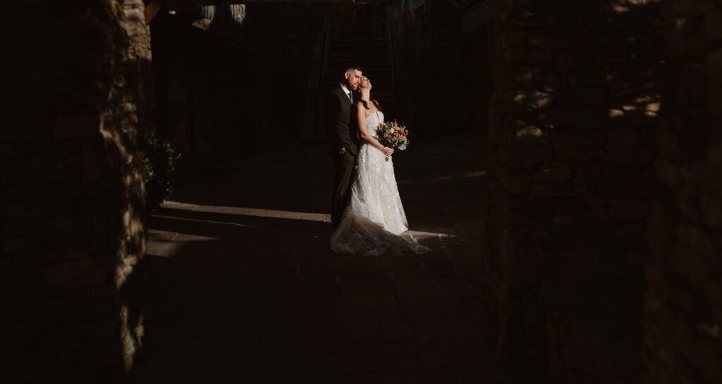 A newlywed couple standing in the sunlight with one partner hugging from behind. 