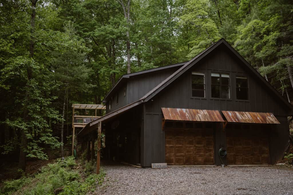 A view of a cabin in the forest with a black exterior and the small parking area.