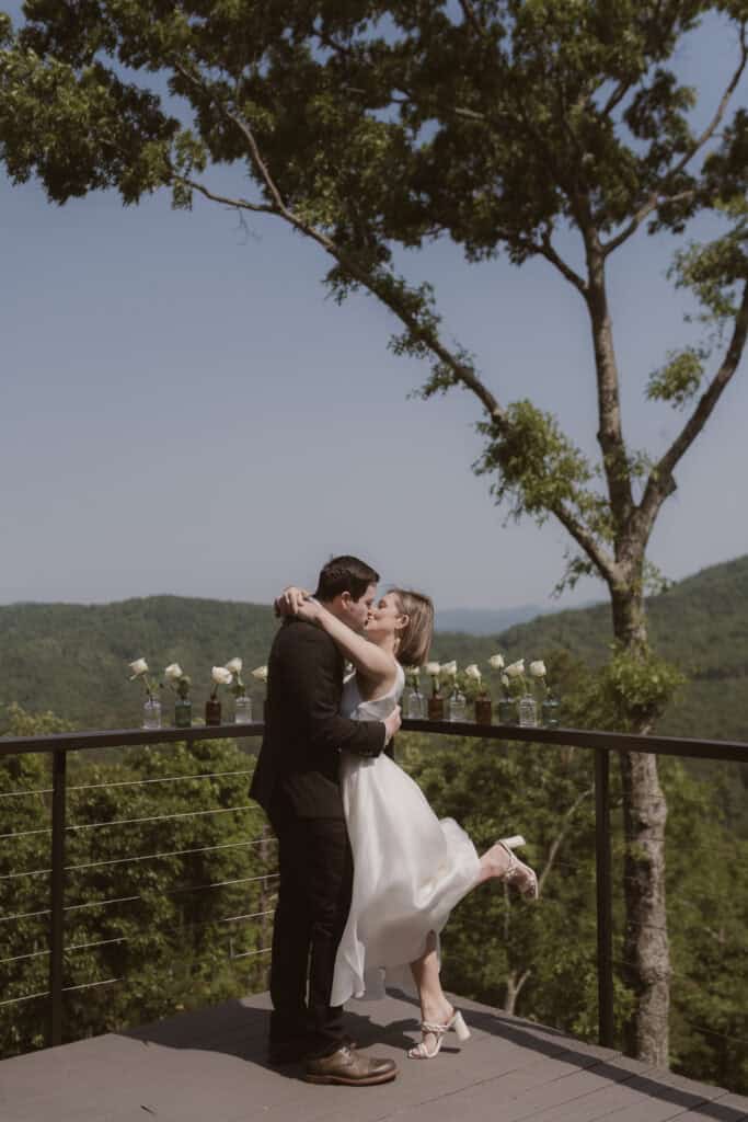 A newlywed couple enjoying their first kiss as a married couple on a large porch with mountains in the distance. 